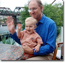 Gabriel siting at table with toddler in lap smiling and waving at camera